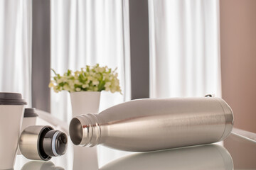 One vacuum insulated metal bottle for water or coffee and cups with coffee on the kitchen table.
