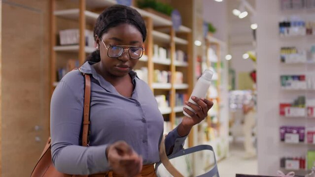 Medium shot of African American woman standing in front of counter in cosmetics store, looking at goods on display, picking up bottles of skincare products, looking at label and putting in basket