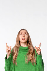 Portrait of surprised woman pointing fingers up, showing advertisement on top, recommending click on link, demonstrating promo banner, wearing green cozy sweater, standing over white background