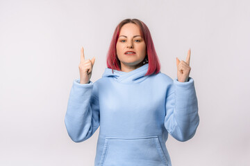 Sad, reluctant young woman with pink hair pointing fingers up and looking upset, complaining at something on top, standing in blue hoodie over white background
