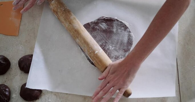 Top view of a table on which lies white baking paper and on it chocolate dough, which a young woman pastry chef rolls out using a rolling pin. The process of preparing cake layers in production.