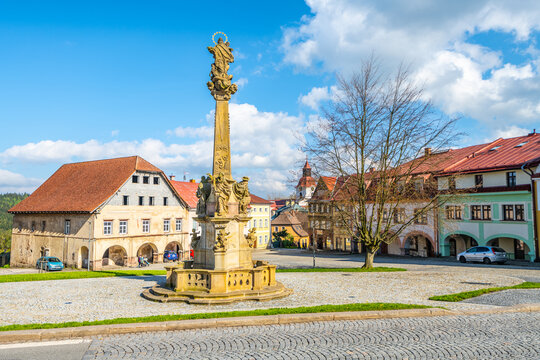 A baroque-style Marian column stands as a centerpiece in Rychorske Square in Zacler, Czech Republic. The square is surrounded by classic European buildings under a bright blue sky.