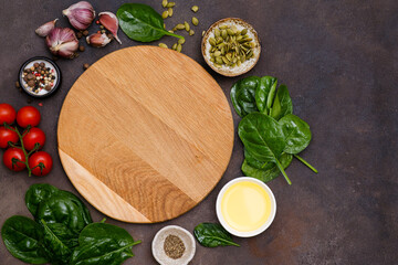 Round wooden cutting board with food ingridients tomatoes, spinach, oil, pumpkin seed, garlic, pepper on brown background, top view,