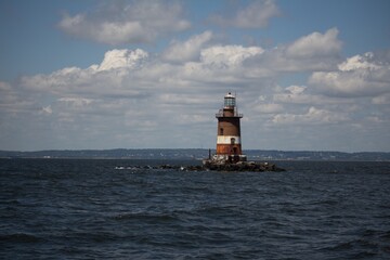lighthouse on small rocky island in the middle of an ocean, distant city skyline visible in background, sunny day with clouds in sky, photograph taken from boat at distance, lighthouses have white and