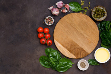 Round wooden cutting board with food ingridients tomatoes, spinach, oil, pumpkin seed, garlic, pepper on brown background, top view,