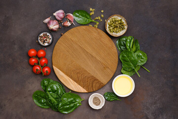 Round wooden cutting board with food ingridients tomatoes, spinach, oil, pumpkin seed, garlic, pepper on brown background, top view,