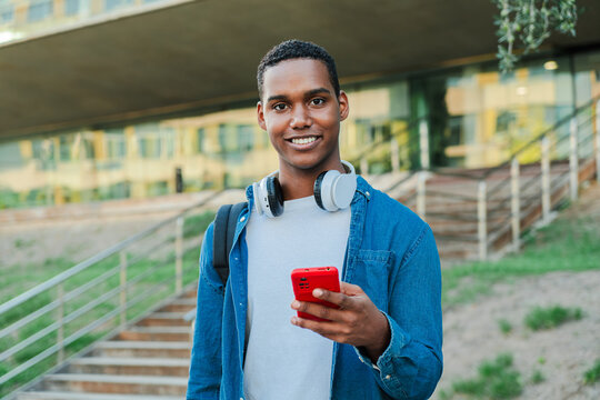 Individual portrait of one african american guy looking at camera and holding a smartphone. Real teenage high school student using a cellphone and staring front at university campus. Man with phone