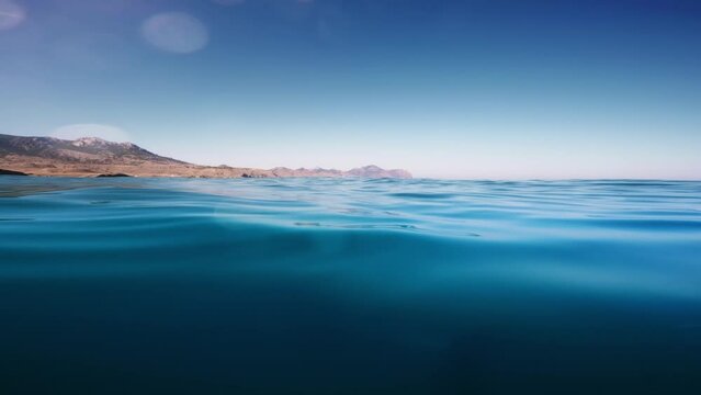 Abstract video background of the ocean with the underwater part and the sun shining above, divided by the waterline. Beautiful clear sky with a mountain range and bright sun over sea water