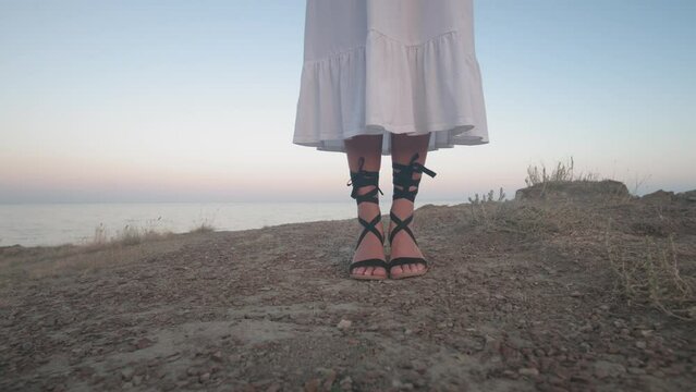 young sexy slender woman posing on an island near the sea in a white dress and black sandals with ties. Greek style of clothing on a beach holiday