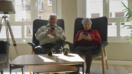 Elderly man and woman couple sitting in armchairs and watching mobile phones