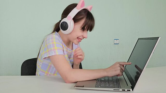 Portrait of little girl in wireless headset using laptop, studying online at home, interested happy student typing on keyboard looking at pc screen