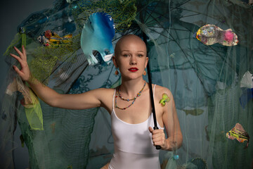 A bald woman in white poses confidently with an underwater-themed set complete with artificial fish and netting