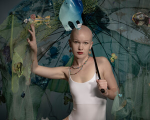 A bald woman poses amid a fantastical underwater scene with fish and a net, conveying a surreal, dreamlike quality
