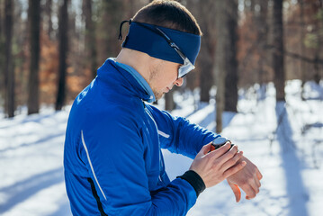 Runner Checking Smartwatch During Winter Training in Snowy Forest. A runner man in a blue jacket and headband pauses to check his smartwatch during a winter training session in a snowy forest setting.