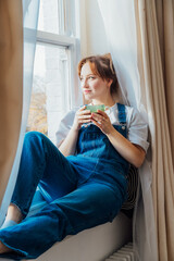 Time To Relax. Young woman looks out the window overlooking the city, sits on windowsill at cozy home holds cup of hot tea drink. Happy calm female taking break for mental health wellbeing. Vertical - obrazy, fototapety, plakaty