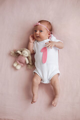 girl baby infant with a bow lies on the bed in a white bodysuit with a doll toy next to it, the child has a pink number 1 months on a pink background