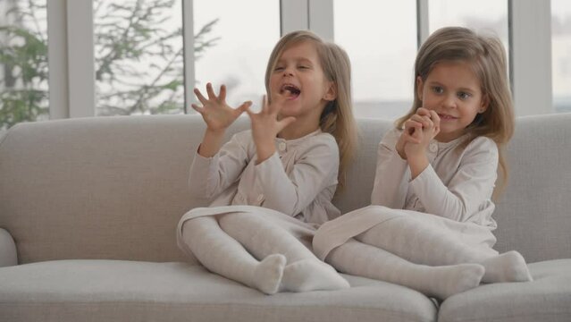 Two children twins playing at home. Happy cheerful mischievous little sisters twins sitting on sofa, smiling and laugh, playing in living room at home. Family having fun, sisters togetherness concept.