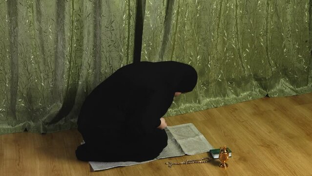 A Muslim woman in a black khimar reads prayer on her knees on a rug at home