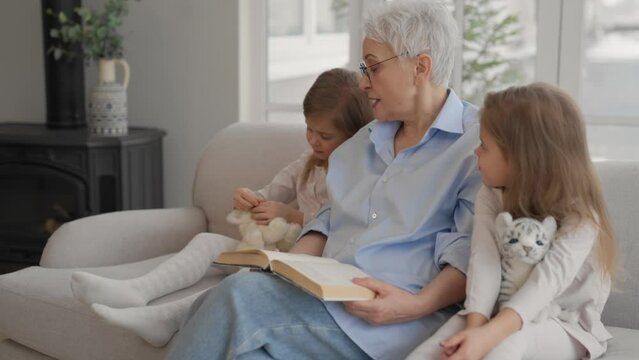 Grandmother with granddaughters resting. Happy relaxed older adult granny embracing reading book with children two twins granddaughters leisure rest on couch at home. Family love, moralizing concept.