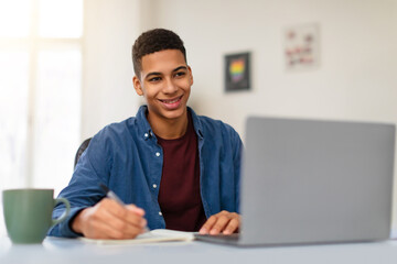 Smiling black teenage guy studying with laptop at home