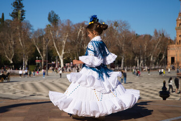 Fototapeta premium a little girl dancing flamenco dressed in a white dress with ruffles and blue fringes in a famous square in seville, spain. The girl has flowers on her head. Flamenco, cultural heritage of humanity.