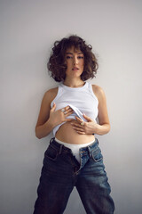 sexy fashionable young curly-haired woman in jeans and white T-shirt standing against the white wall of the house. showing white pants.