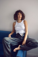 sexy fashionable young curly-haired woman in jeans and white T-shirt sitting on the blue sofa against the white wall of the house. showing white pants.