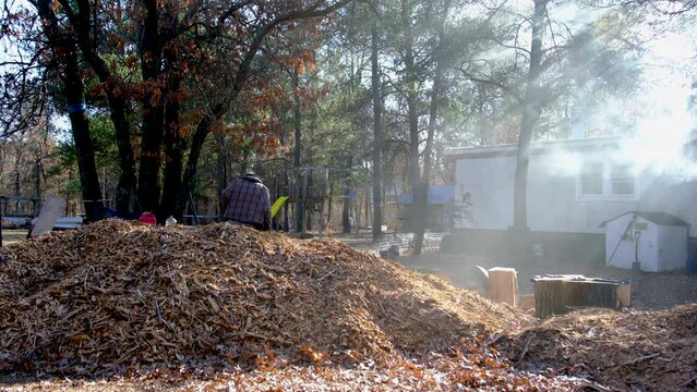 Man in front of pile of mulch using ax to split or process oak lumber into firewood meanwhile smoke rise and wafting around scene from mobile house chimney as wood being burn in cast iron stove or fir