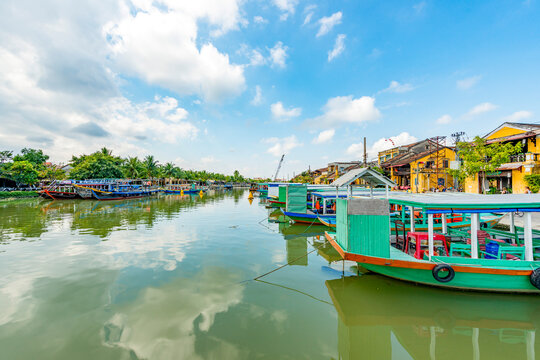 Wooden boats on the Thu Bon River in Hoi An Ancient Town (Hoian), Vietnam. Yellow old houses on waterfront reflected in river. Riverside Scenes in the Historic District of Hoi An, Vietnam