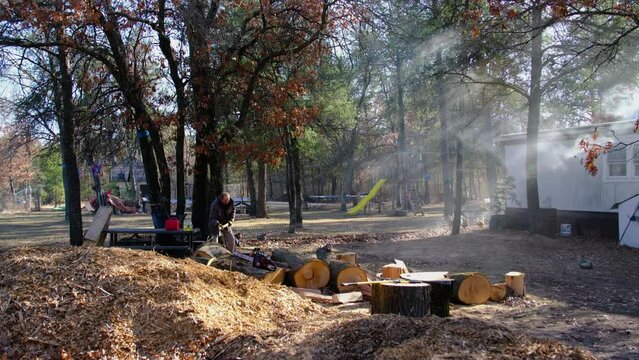 Young adult processing oak lumber into firewood using Russian style ax meanwhile smoke wafting from manufactured house chimney onto scene as wood being burn in fireplace.