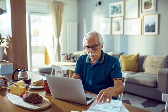 Senior man with glasses reviewing bills and using laptop at home