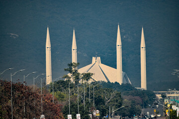 A Faisal mosque with tall minarets, amidst trees and streetlights, against a mountain backdrop...