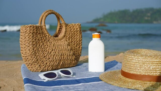 Beach essentials arranged on sandy shore bottle of sunscreen, stylish straw hat, sunnies atop towel, wicker tote aside. Products against UV rays for summer skin protection on tropical coast.