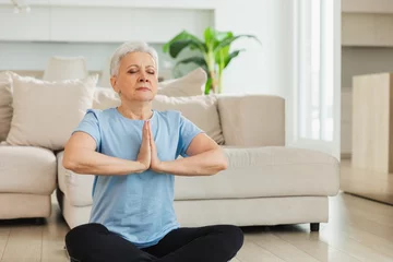Fototapeten Yoga mindfulness meditation. Senior adult mature woman practicing yoga at home. Mid age old lady sitting in lotus pose on yoga mat meditating relaxing. Older middle aged woman doing breathing practice © Юлия Завалишина