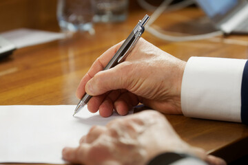 An elderly man in a business suit with cuffs, writing with a fountain pen on a piece of paper....