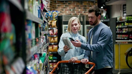Young caucasian husband standing near shopping cart while attractive wife taking package from shelves in grocery store. Couple dressed in casual attire. Aisle with snacks, colorful bags and boxes. - Powered by Adobe