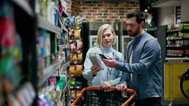 Young caucasian husband standing near shopping cart while attractive wife taking package from shelves in grocery store. Couple dressed in casual attire. Aisle with snacks, colorful bags and boxes.
