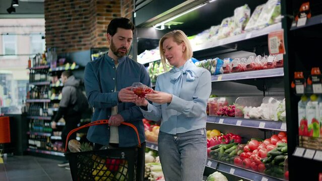 Lovely married couple examining product in produce section while standing near shelves stocked with fresh fruits and vegetables. Husband holding basket filled with various items and waiting for wife.