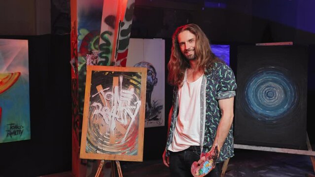 Portrait of smiling long haired artist holding paint palette and standing near picture on easel in colorful art studio. Original caucasian male presenting creative work indoors.