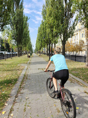 Naklejka premium A young blonde woman rides a bicycle along the alley in perspective. Rear view. Tourism, sports and outdoor recreation