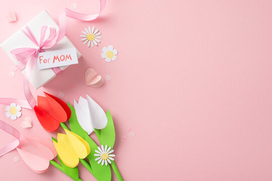 DIY Mother's Day concept, top view snapshot of paper tulips, chamomiles, a self-made gift box inscribed "for mom", ribbon-tied, amidst paper hearts and fine confetti on a pastel pink canvas