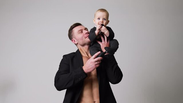 Handsome man wearing a black jacket on the naked torso holding a baby on his shoulder. Father and infant son portrait. Studio footage. White backdrop.
