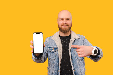 Smiling bald bearded man is pointing at a phone with blank screen he holds while standing over...