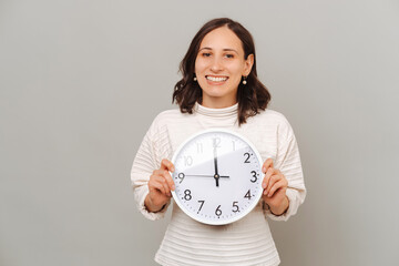 Studio portrait of a cheerful young woman holding a round clock over grey backdrop.