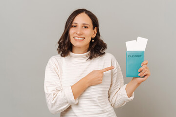 Studio portrait of a cheerful woman pointing at the passport she holds over grey background.