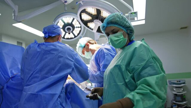 Female scrub nurse wearing protective uniform and mask passes tools to the chief surgeons. Trauma surgery progress in modern operational room.
