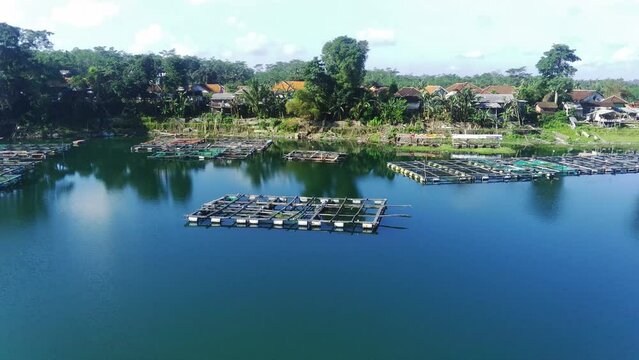 drone video captures a scenic village nestled on the shores of Ranu Pakis lake in Lumajang, East Java, Indonesia. Lush greenery surrounds the quaint houses with colorful roofs. Smoke curls from chimne
