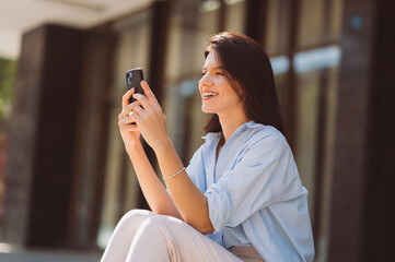 Cheerful young brunette woman is using phone while sitting outdoors on a sunny day.