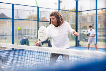 Portrait of active concentrated man playing padel tennis on open court on warm autumn day, swinging racket to return ball over net .. - obrazy, fototapety, plakaty