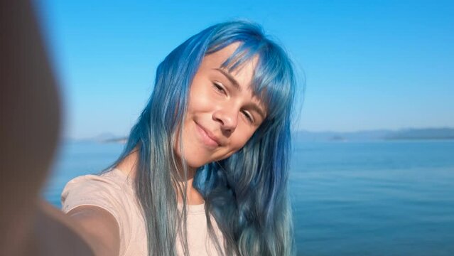 Blue haired teen make summer selfie. A young blue haired girl hold her phone and makes traveling video from the sunny seashore in summer.